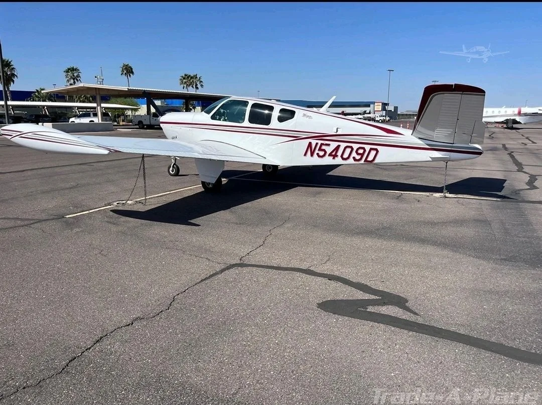 A white Beechcraft Bonanza (N5409D) with red stripes is parked on an airport tarmac under a clear blue sky.