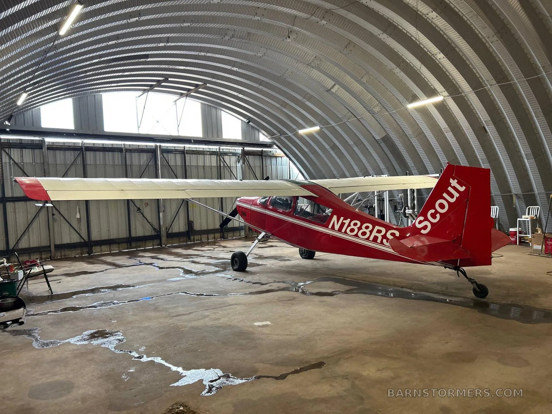 A red Scout N188RS aircraft with white stripes is parked inside a metal hangar, viewed from the rear-right.