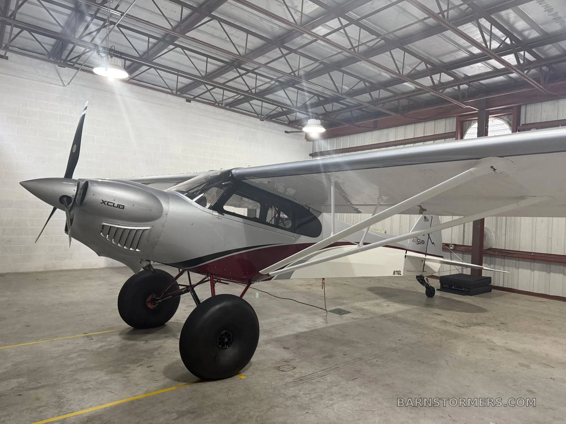 A silver and red X-Cub aircraft with large tundra tires, parked inside a hangar, viewed from the front-left.
