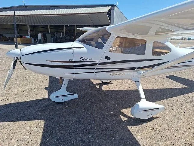 A close-up side view of the front of a white Sirius LightSport aircraft with black and grey stripes, showing the propeller and nose.