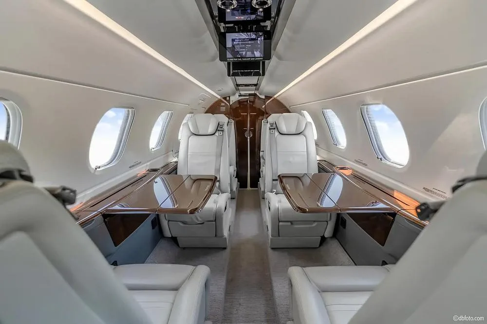 A view of the private jet's cabin interior, showing rows of light-colored seats with pull-out tables and overhead lighting.