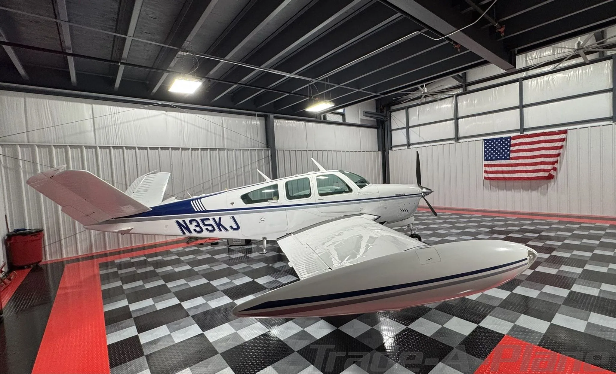 A white Beechcraft Bonanza N35KJ with blue and grey stripes parked in a hangar with an American flag on the wall.