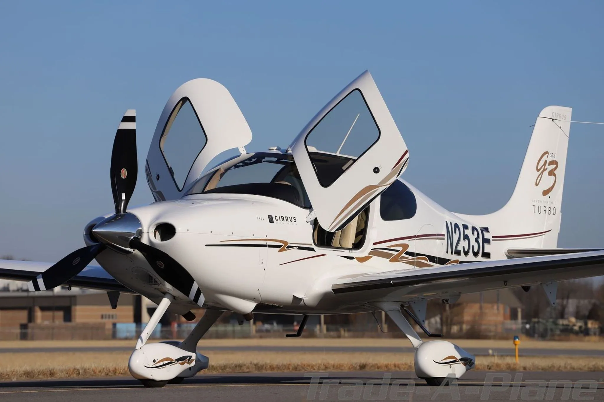 Side-front view of a white Cirrus SR22T aircraft (N253E) with its doors open and gold and brown stripe livery, parked on an airfield.