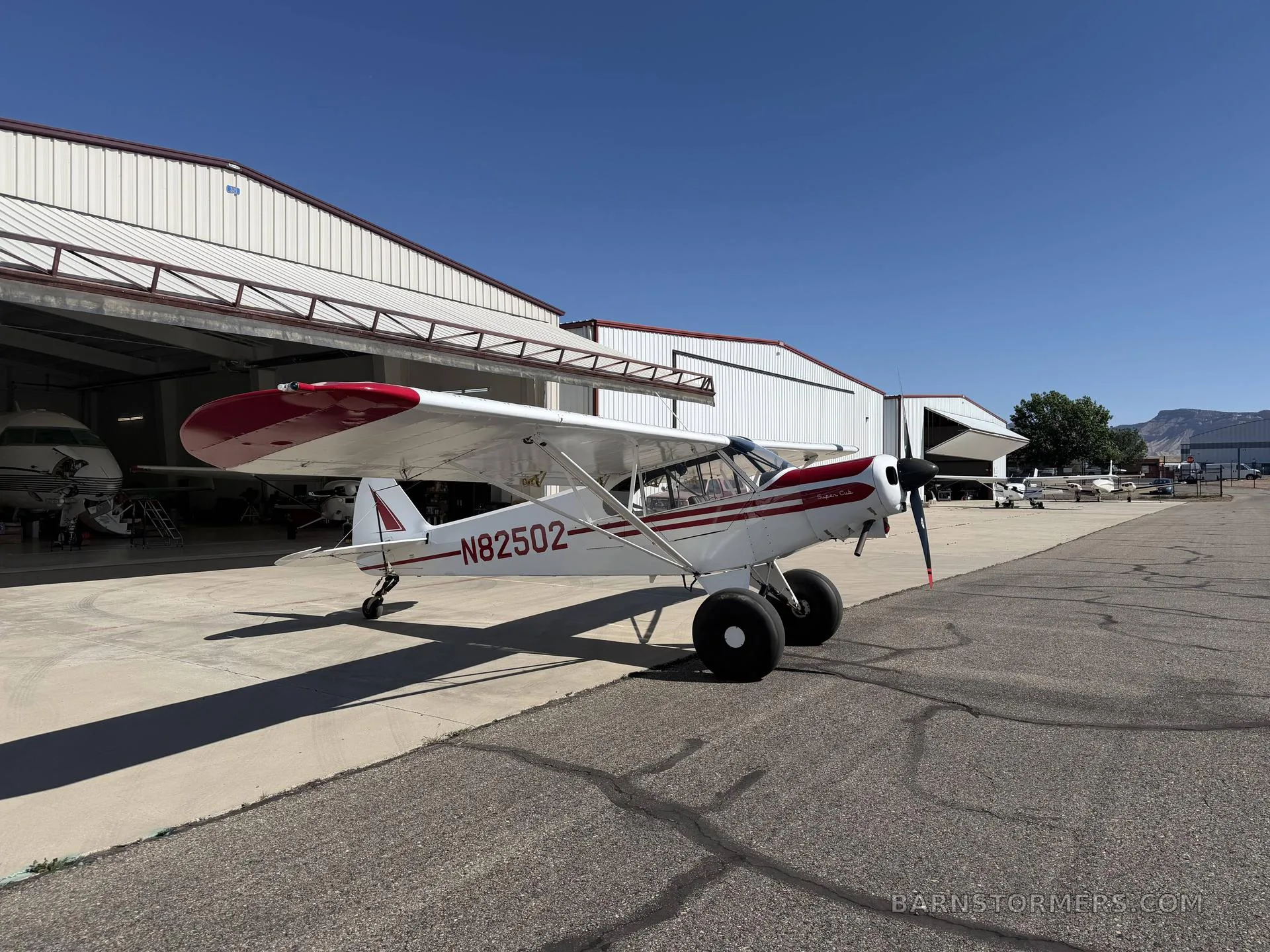 A white and red single-engine bush plane with large tires is parked on an asphalt apron in front of hangars under a clear blue sky.