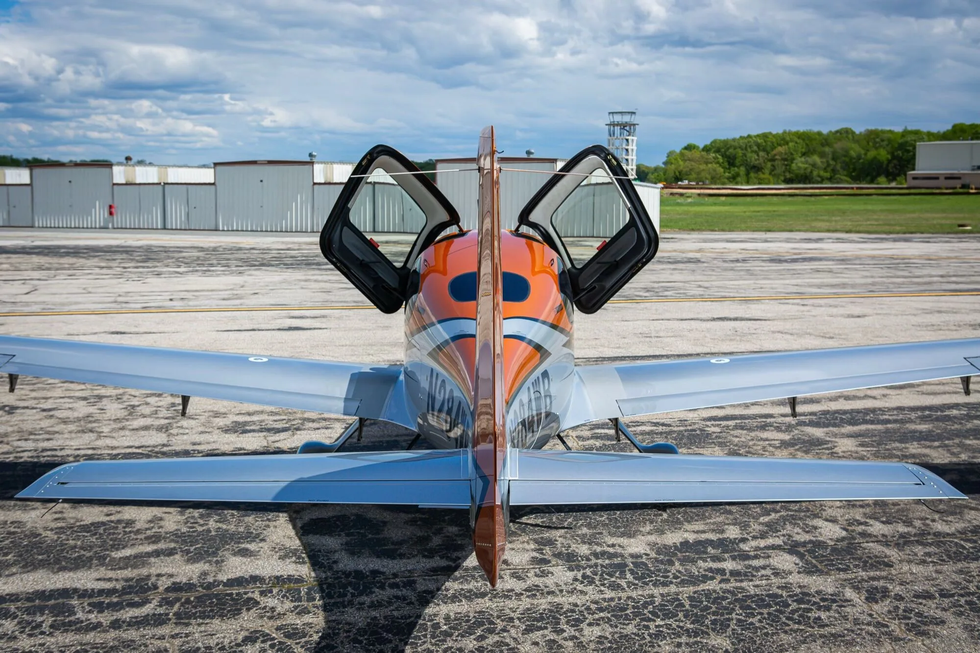 A rear view of an orange and silver Cirrus SR20 aircraft with its gull-wing doors open, parked on an airfield.