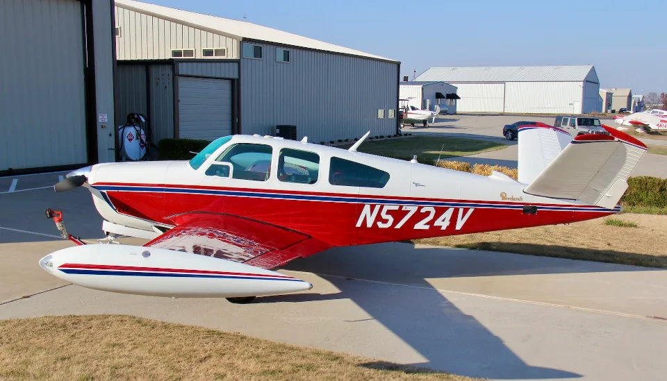 A three-quarter front view of a red and white Beechcraft Bonanza N5724V parked in front of hangars.