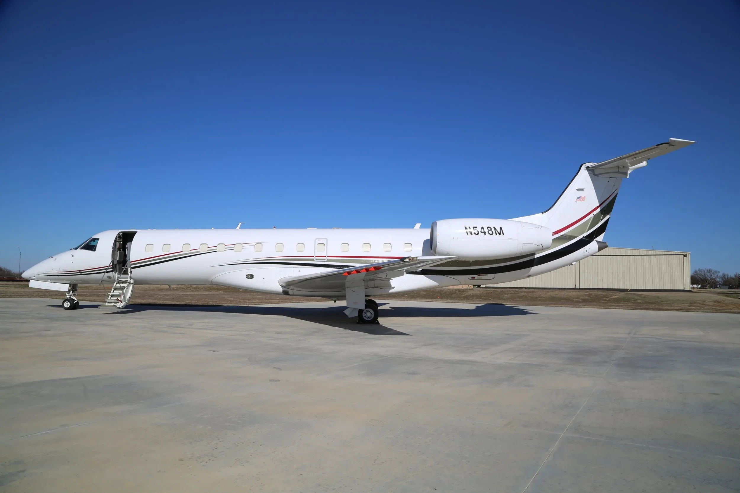 A white Embraer Legacy business jet with its main cabin door open and stairs extended is parked on a tarmac.