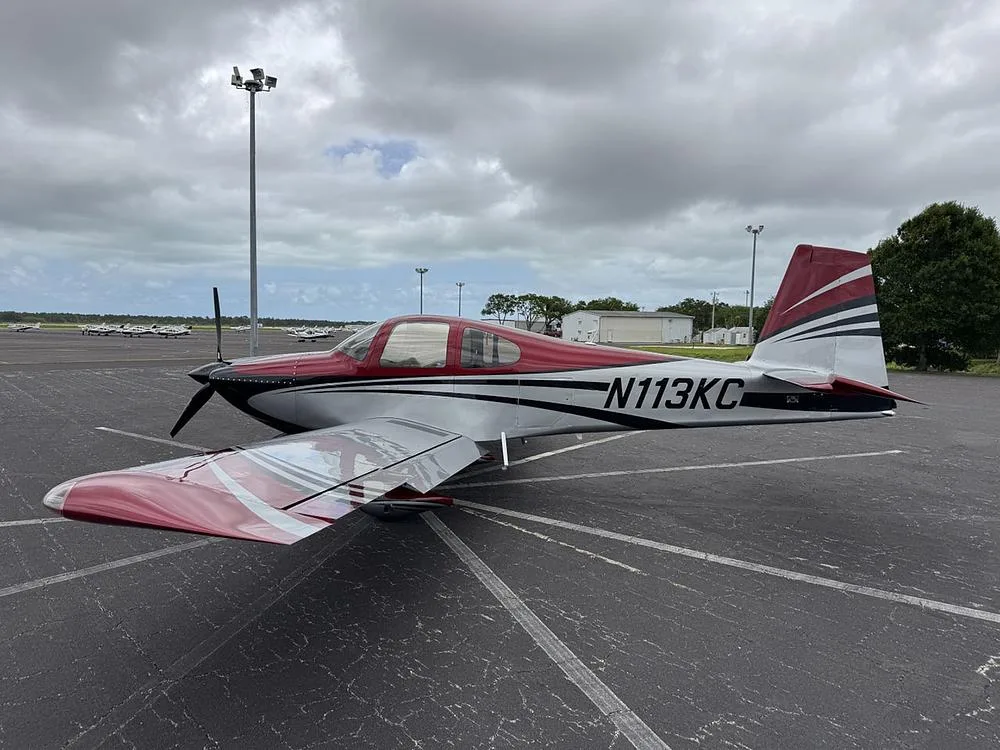 A red, silver, and black single-engine aircraft (N113KC) parked on an airport tarmac under an overcast sky.