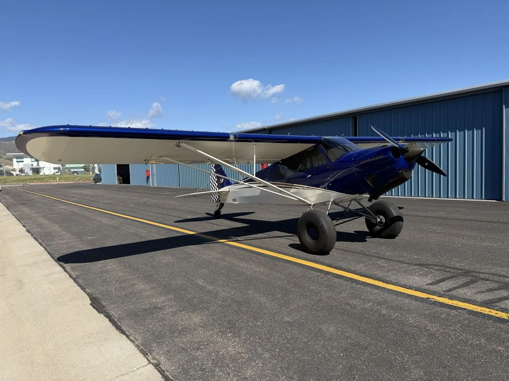 A blue and white Carbon Cub SS aircraft with large tundra tires, seen from the front-left side on an airport tarmac.