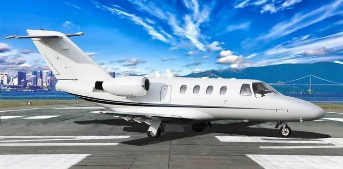 A white private jet, likely a Cessna Citation, on a runway with a city skyline and bridge in the background.