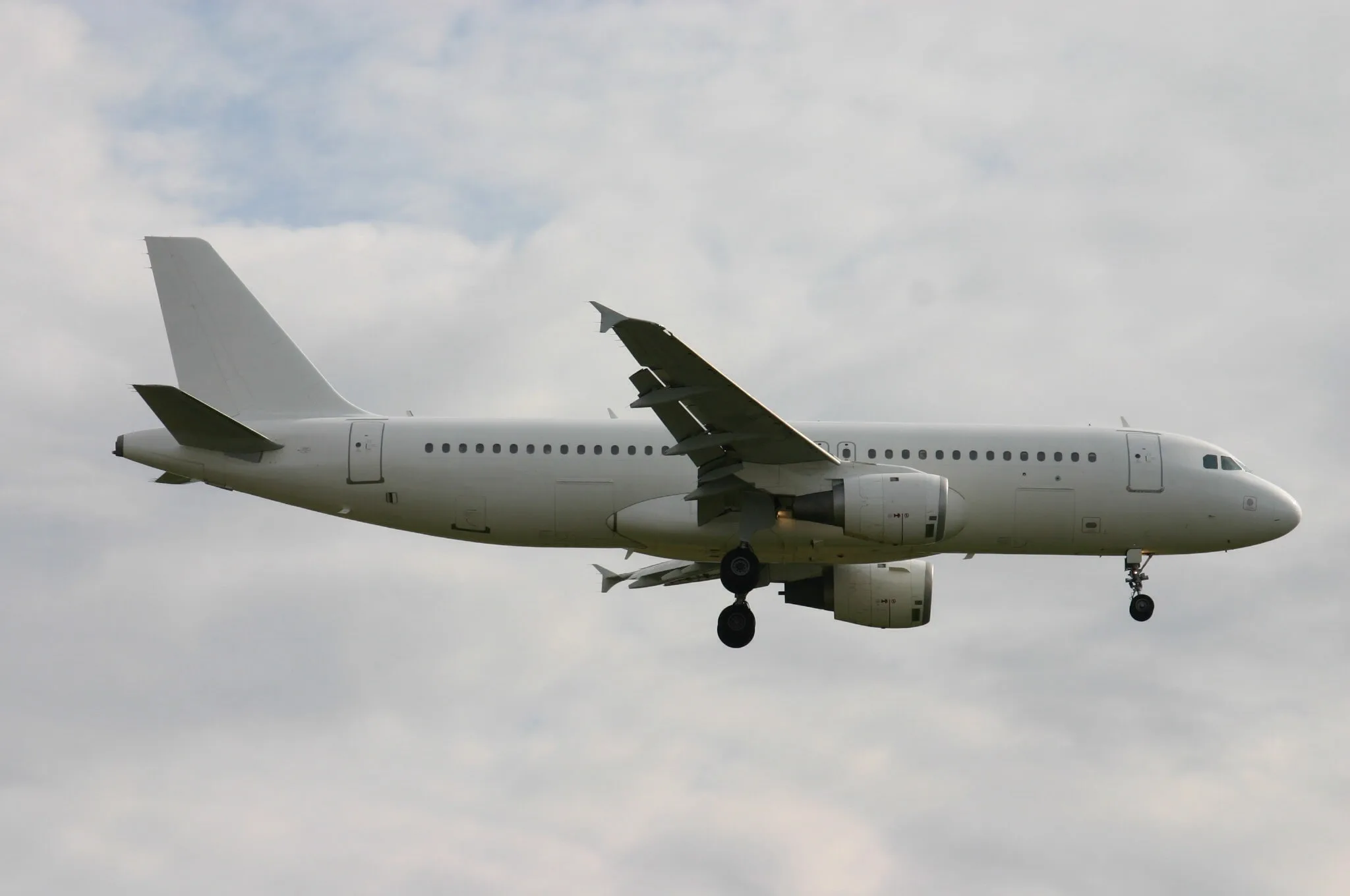 A white, unpainted narrow-body airliner with landing gear extended flies against a cloudy sky.