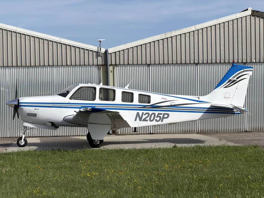 A white and blue Beechcraft Bonanza N205P parked on a paved surface in front of a corrugated metal hangar.