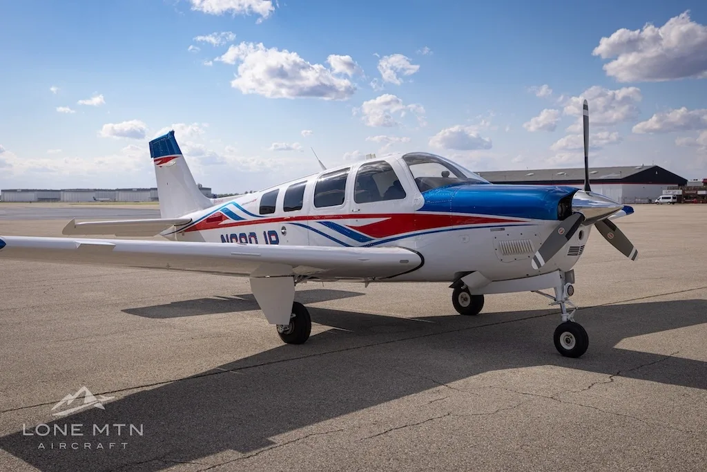 A white, blue, and red Beechcraft Bonanza N680JP parked on an airfield under a partly cloudy sky.