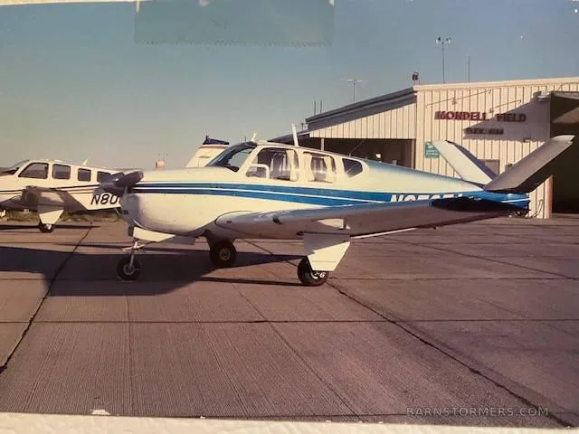 A white Beechcraft Bonanza with blue stripes is parked on a tarmac at Mondell Field, with another aircraft and a hangar in the background.