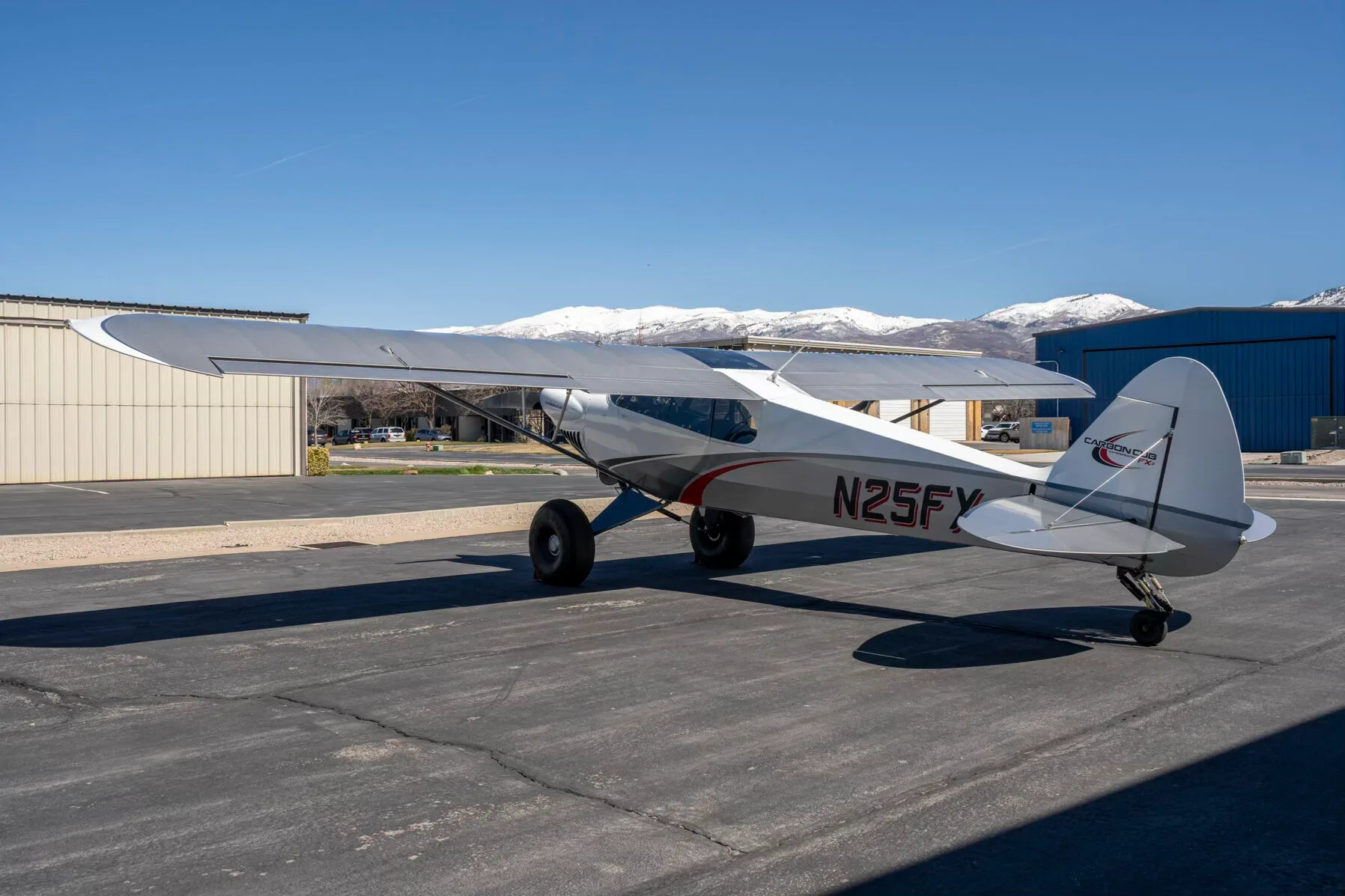 A three-quarter rear view of the white and grey Carbon Cub FX-3 aircraft N25FX parked on an asphalt ramp with snowy mountains in the background.