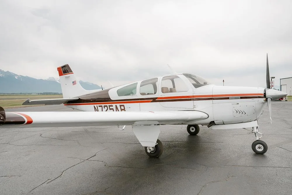 A white single-engine Beechcraft Bonanza aircraft with brown and orange stripes parked on a tarmac, with mountains in the background.