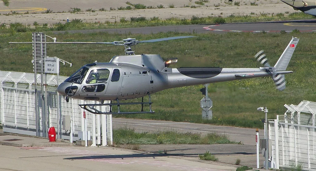 A silver Airbus H125 helicopter with a Turkish flag on its tail, hovering near a fence at an airfield.