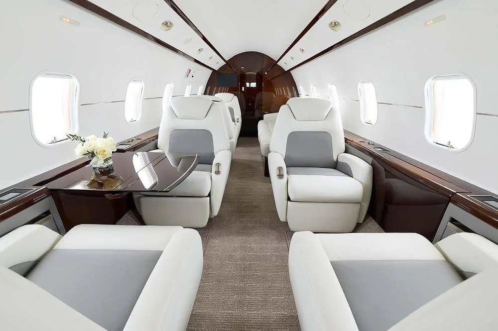 The luxurious interior cabin of a business jet, featuring white and grey leather seats, a dark wood table, and a floral arrangement.