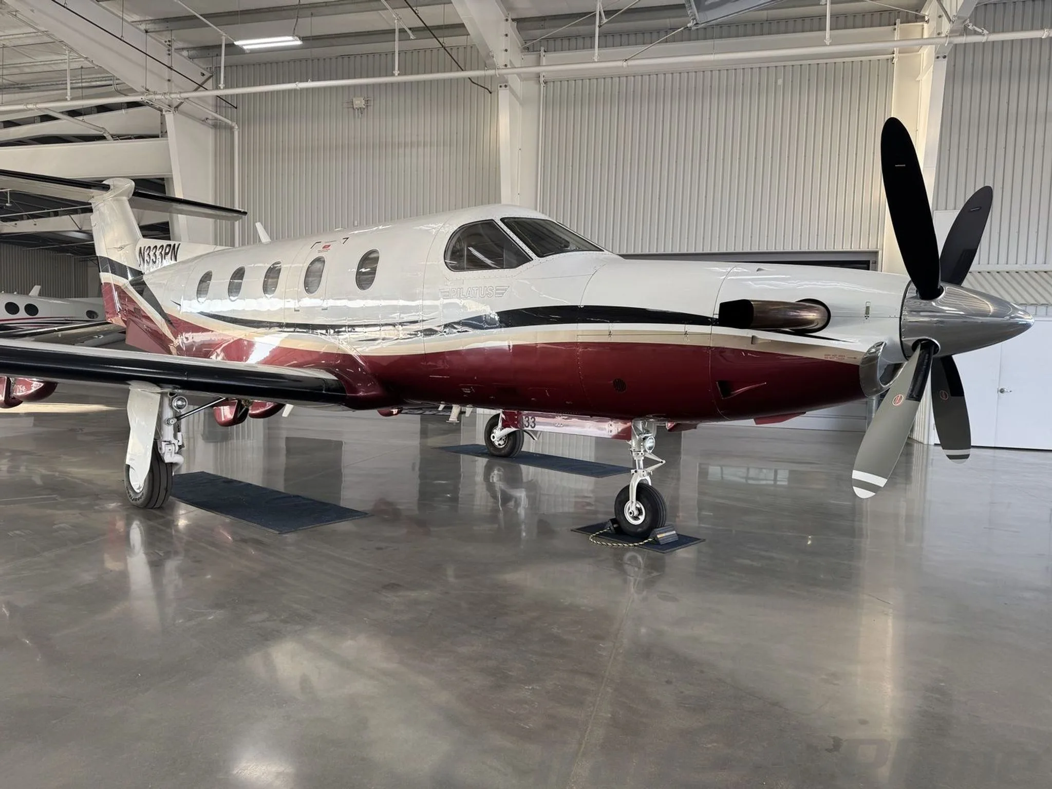 A Pilatus PC-12 turboprop aircraft with a white and maroon paint scheme, registration N333PN, viewed from the front-right in a hangar.