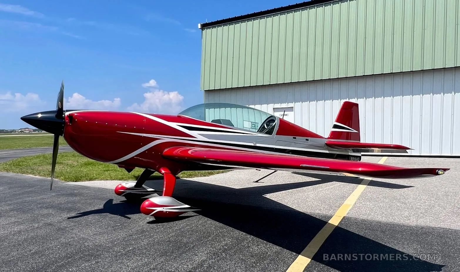 A red and silver aerobatic aircraft with a black propeller is parked on an asphalt tarmac next to a green hangar.