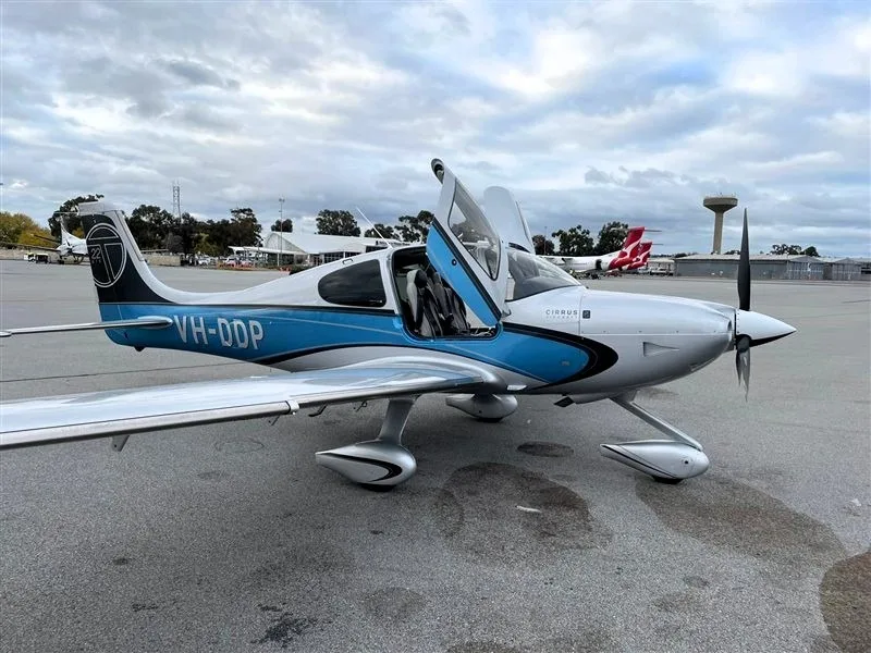 A silver and blue Cirrus SR22T aircraft with open doors parked on an asphalt ramp under a cloudy sky.