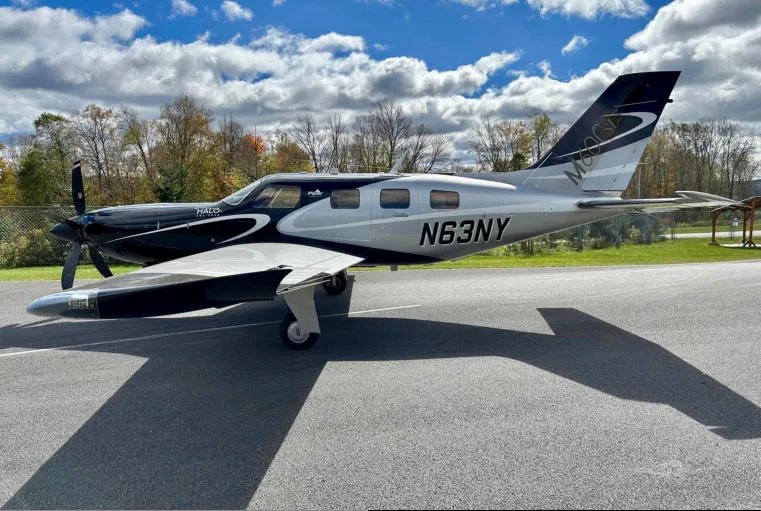 A black and silver Piper M600 SLS aircraft (N63NY) parked on an asphalt runway with autumn trees in the background.