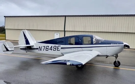 A white and blue Bellanca 7GCBC Citabria N7649E parked on a wet tarmac next to a hangar.