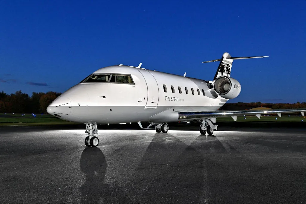 A front-quarter view of a white Bombardier Challenger 605 business jet, registration N605JE, on a tarmac at night.