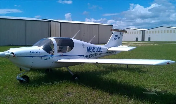 A white Liberty XL2 aircraft with blue stripes and registration N553XL is parked on a grassy field with hangars in the background.