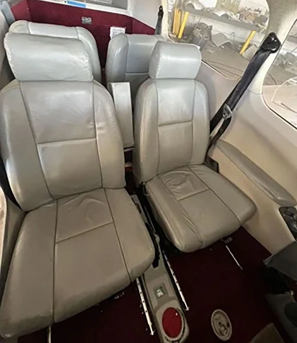 The interior cabin of a small aircraft features two rows of light gray leather seats with a maroon carpet.