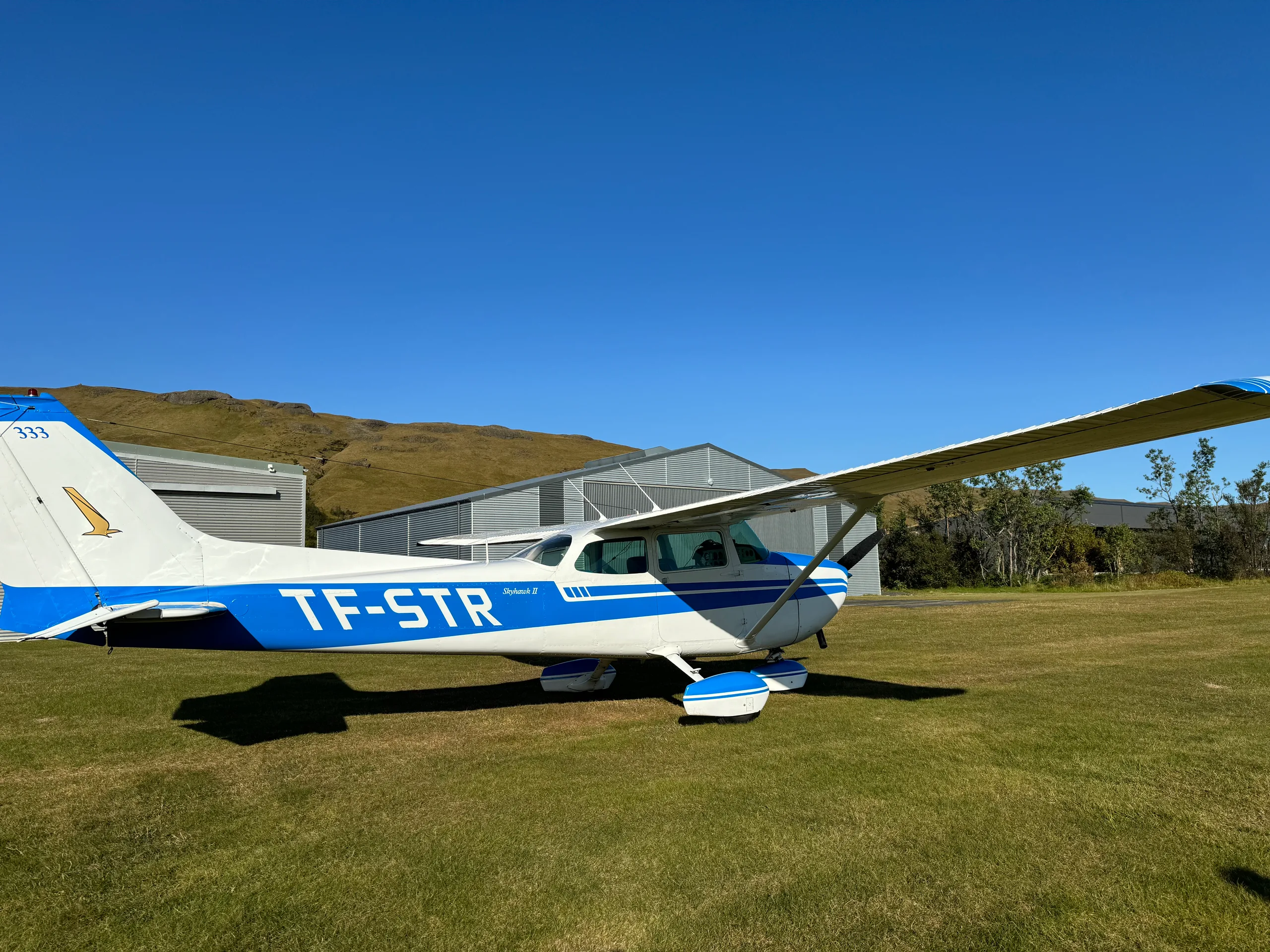 A white and blue Cessna Skyhawk II (TF-STR) parked on a grassy field with hangars and hills in the background under a clear blue sky.