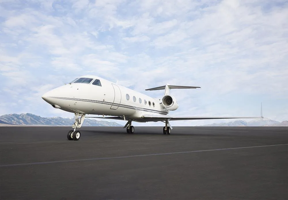 A white Gulfstream G550 business jet with dark blue stripes on a tarmac with mountains in the background.
