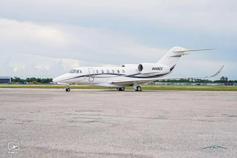 A white Cessna Citation XLS+ business jet with blue and black stripes, tail number N448CX, parked on an airfield under a cloudy sky.