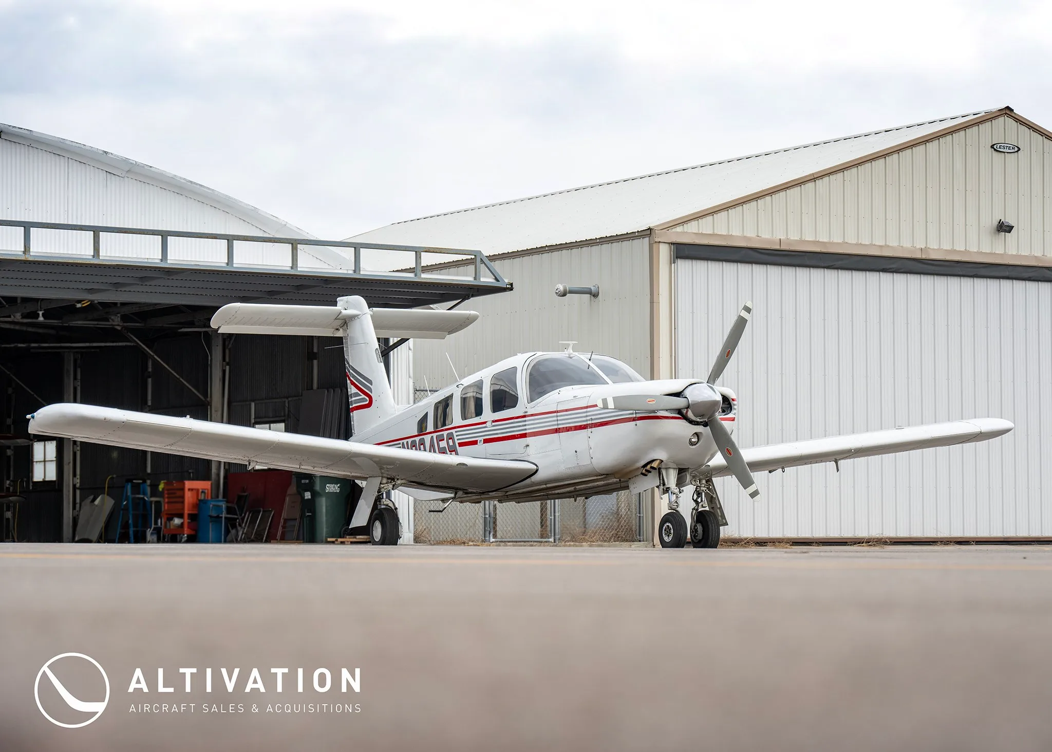 A white single-engine propeller aircraft with red and grey stripes parked on an apron in front of hangars.
