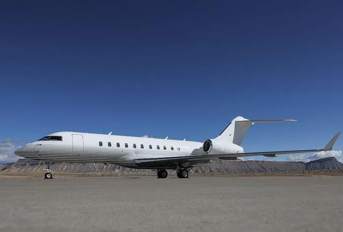 A white business jet with a striped lower fuselage is parked on a tarmac with mountains in the background under a clear blue sky.