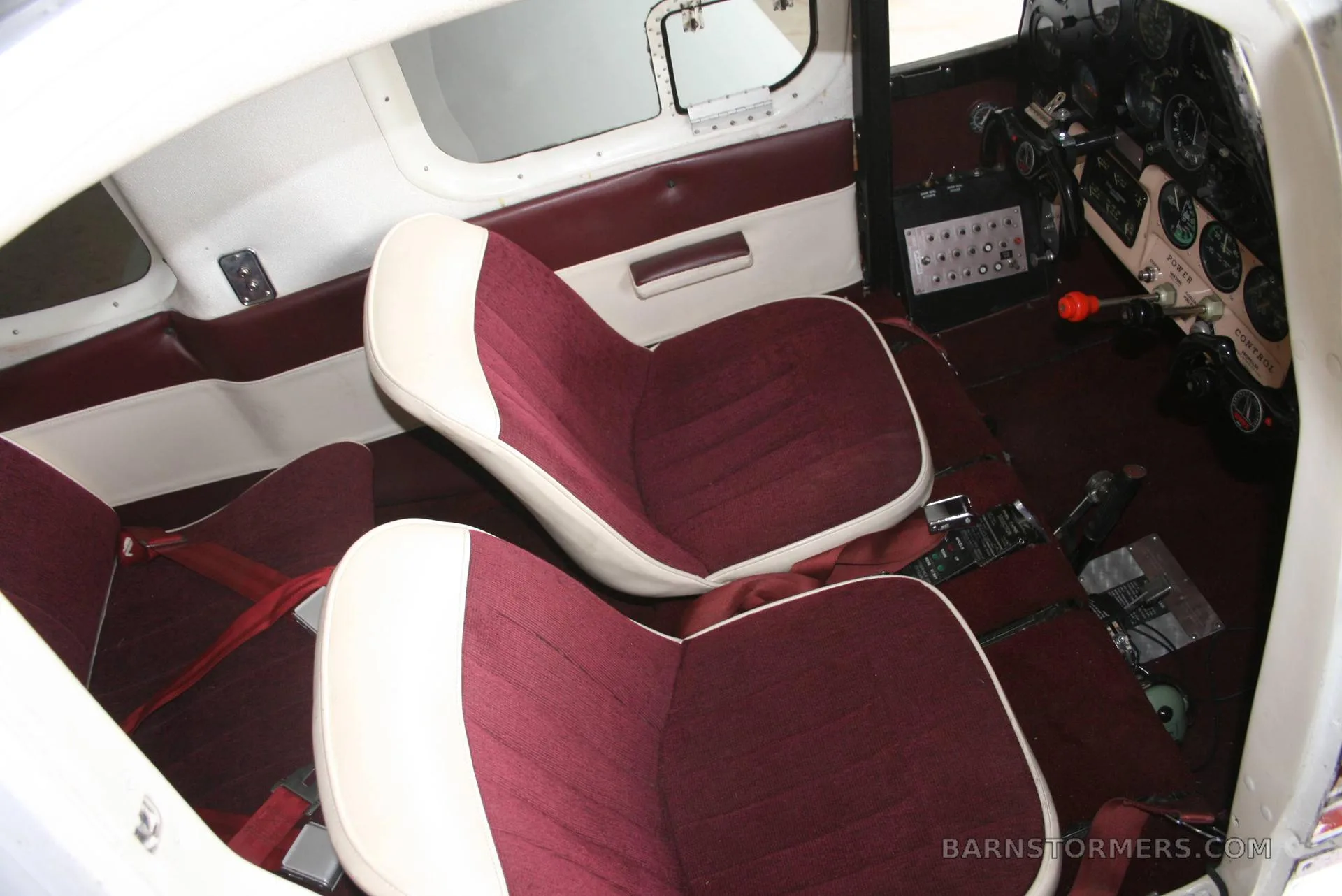 The interior cabin of an aircraft with two maroon and white upholstered seats and a view of the cockpit controls.