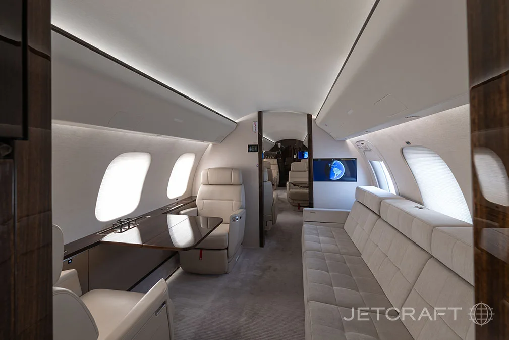 The spacious interior of a business jet cabin features a long beige sofa, individual seats, a table, and a wall-mounted screen displaying a map.