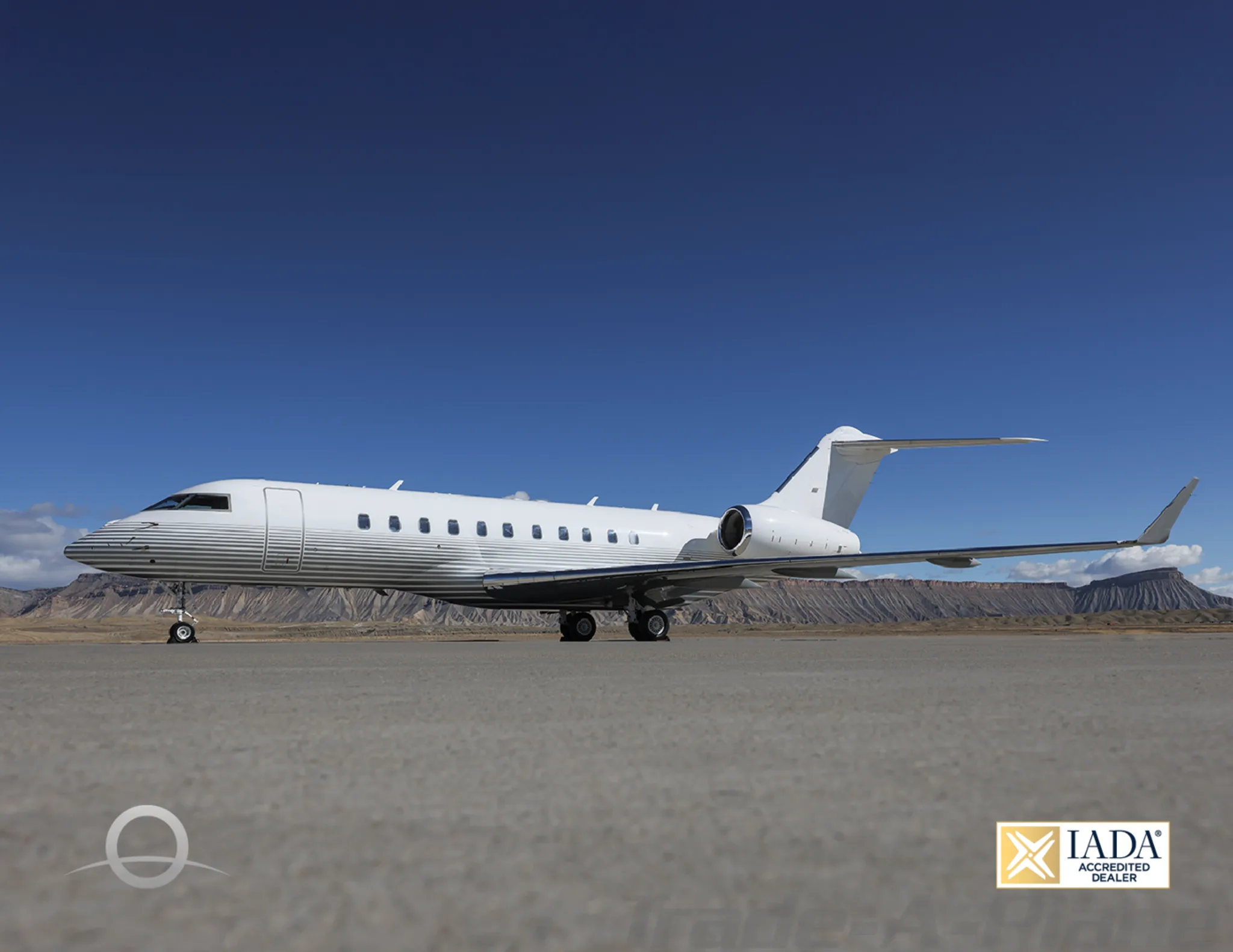 A white Bombardier Global 6000 business jet with grey pinstripes on the fuselage is parked on a tarmac with mountains in the background.