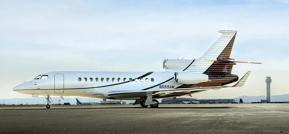 A Dassault Falcon 900EX business jet with white and brown livery is parked on an airfield with an air traffic control tower in the background.