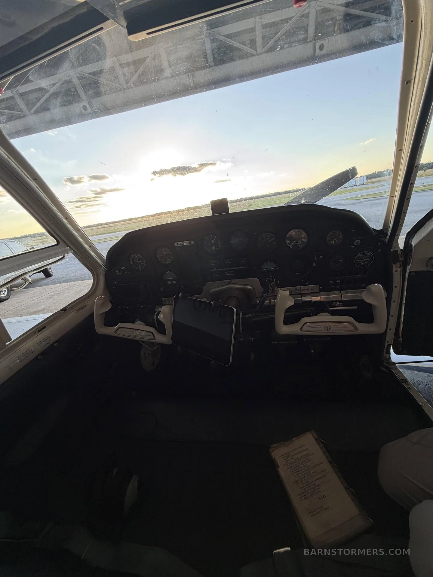 The cockpit of a small aircraft, showing the instrument panel and controls, with a bright sunset visible through the windshield on an airfield.