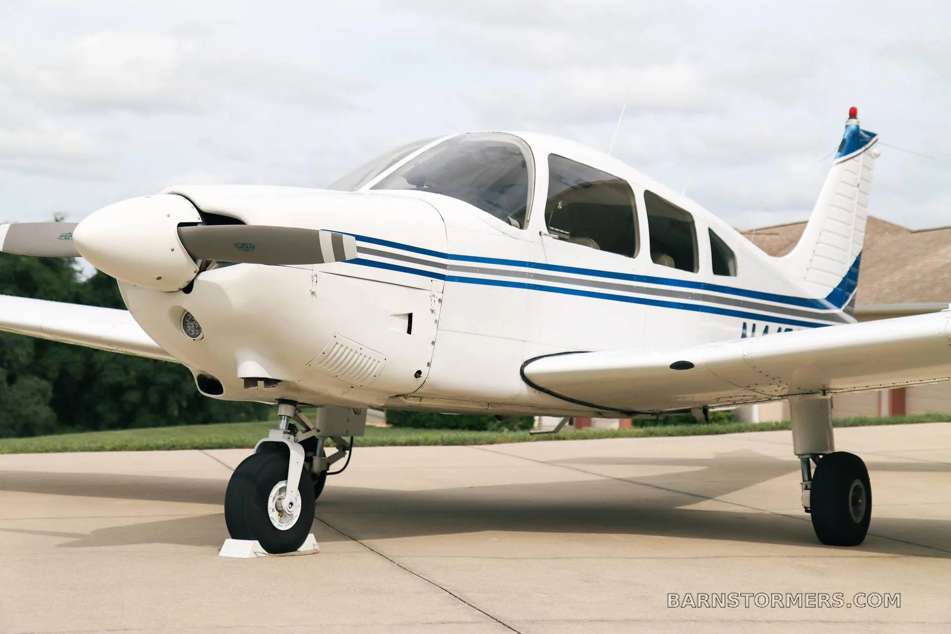 A white Piper PA-28 series aircraft with blue stripes, viewed from the front-left, parked on a concrete pad under a cloudy sky.