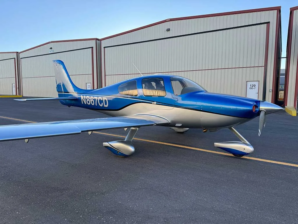 A blue and silver Cirrus SR22 aircraft with registration N867CD parked on an airfield in front of hangars.