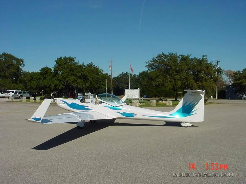 A white motorglider with blue wave decals on the fuselage and tail is parked on an asphalt lot under a clear blue sky.
