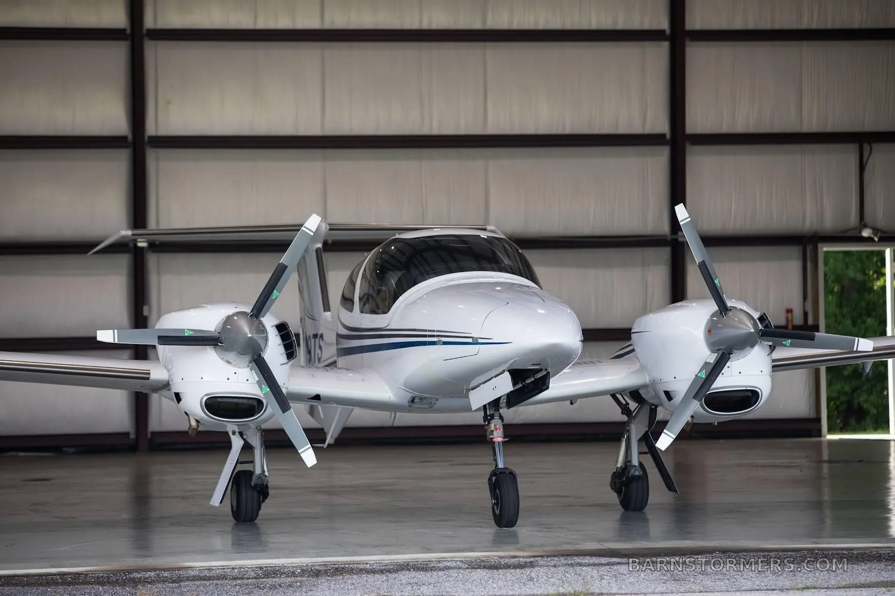 A front view of a white Diamond DA42 aircraft with blue stripes, registration N199TS, parked in a hangar.