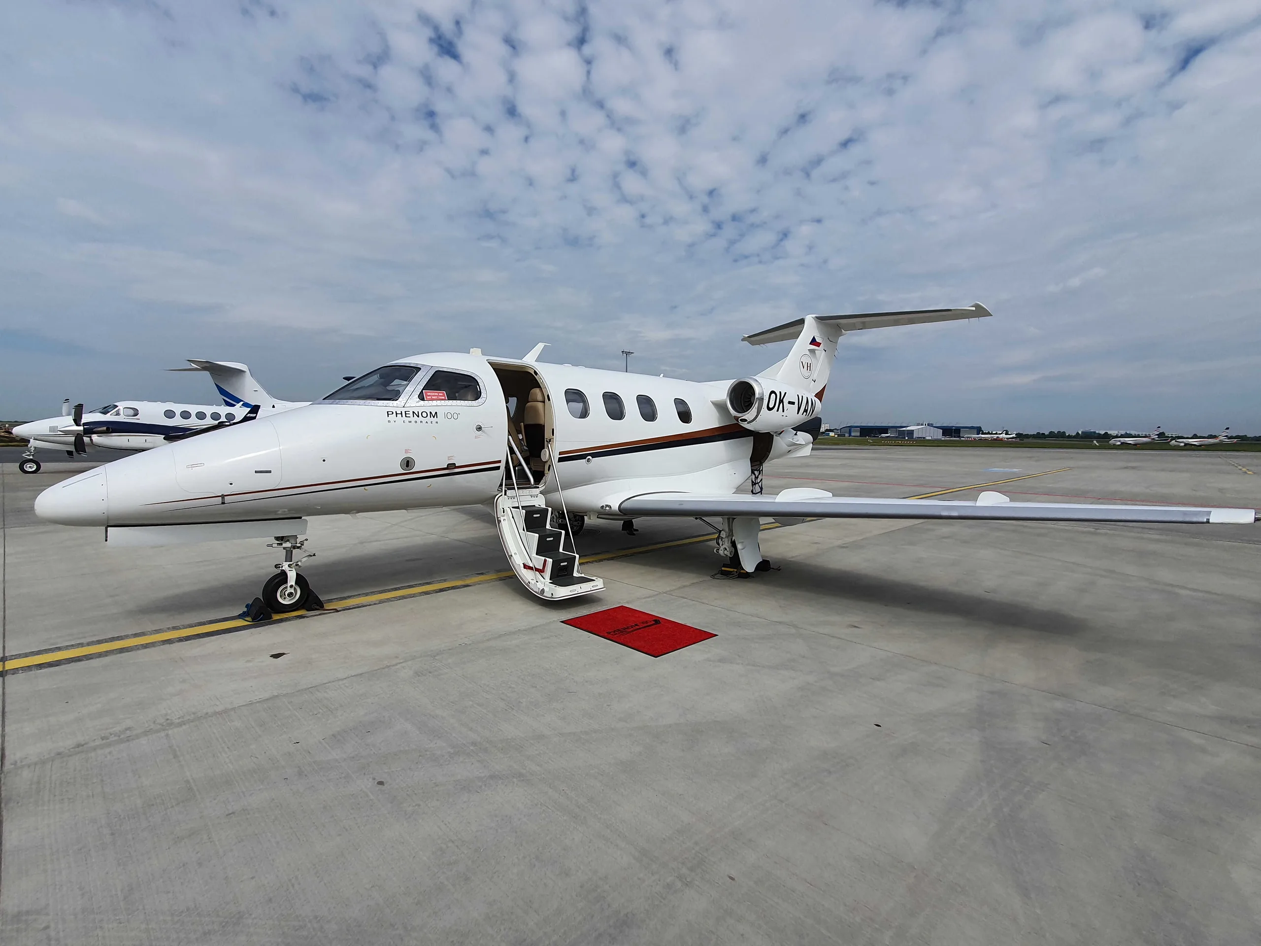 A white Embraer Phenom 100 private jet with brown and black stripes and its door open is parked on an airport tarmac under a cloudy sky.