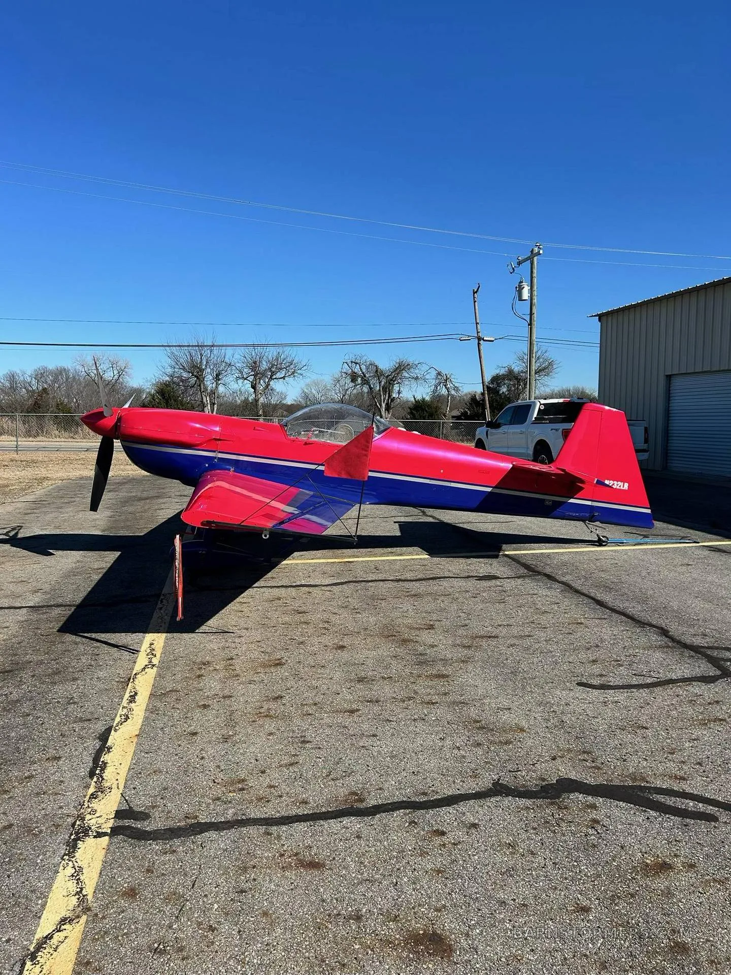 A red and blue aerobatic aircraft, possibly an Extra 300L, is parked on an asphalt apron under a clear blue sky.