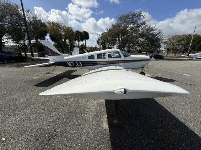 A white Piper Cherokee aircraft with blue and red stripes, registration N7JJ, parked on an asphalt ramp.