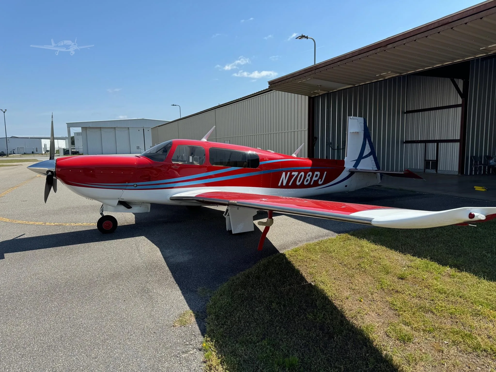 A red and white single-engine propeller aircraft with blue stripes is parked on an asphalt ramp next to a hangar.