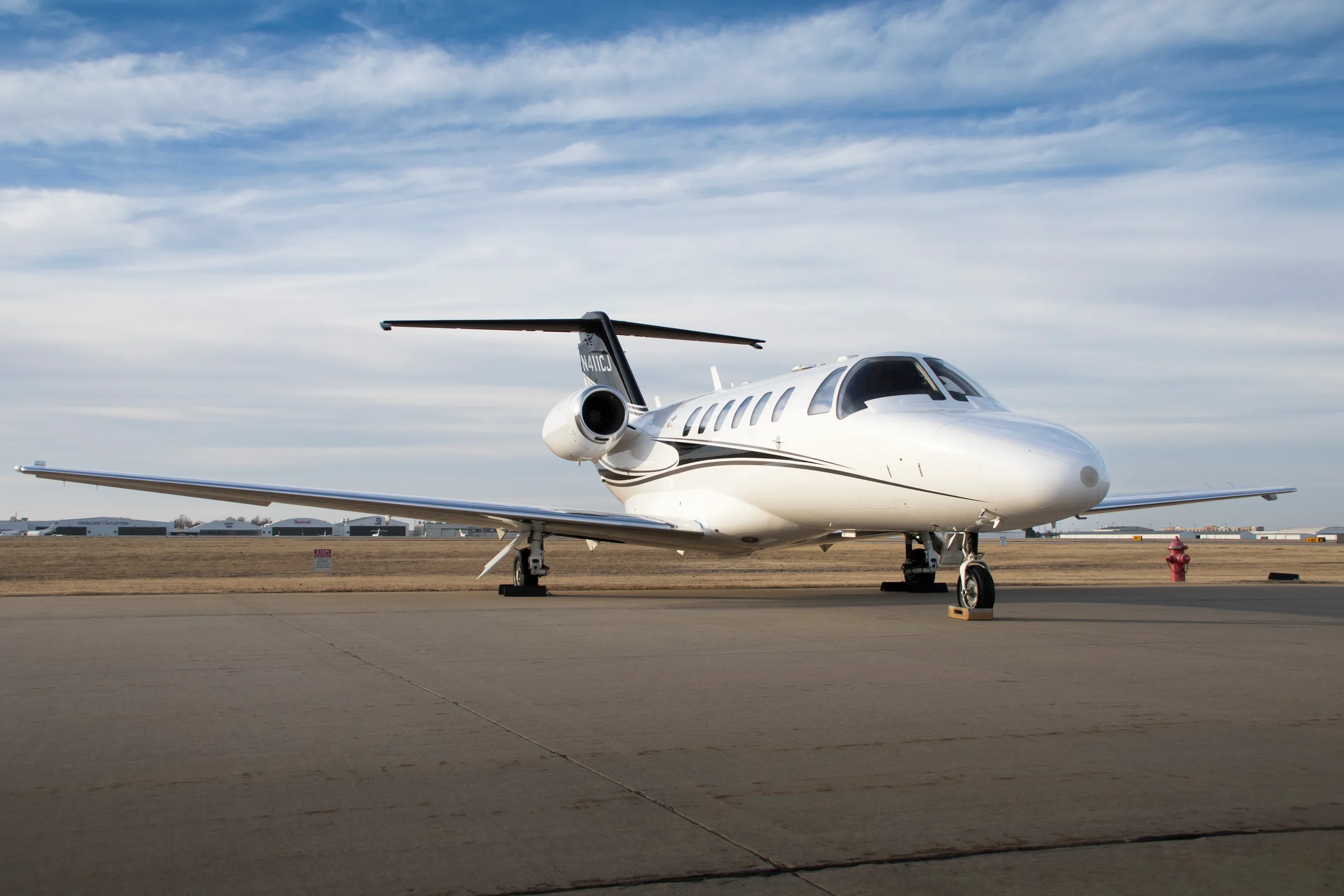 A white Cessna Citation CJ3 private jet with black and gold stripes on the tail, registration N411CJ, parked on an airfield under a partly cloudy sky.