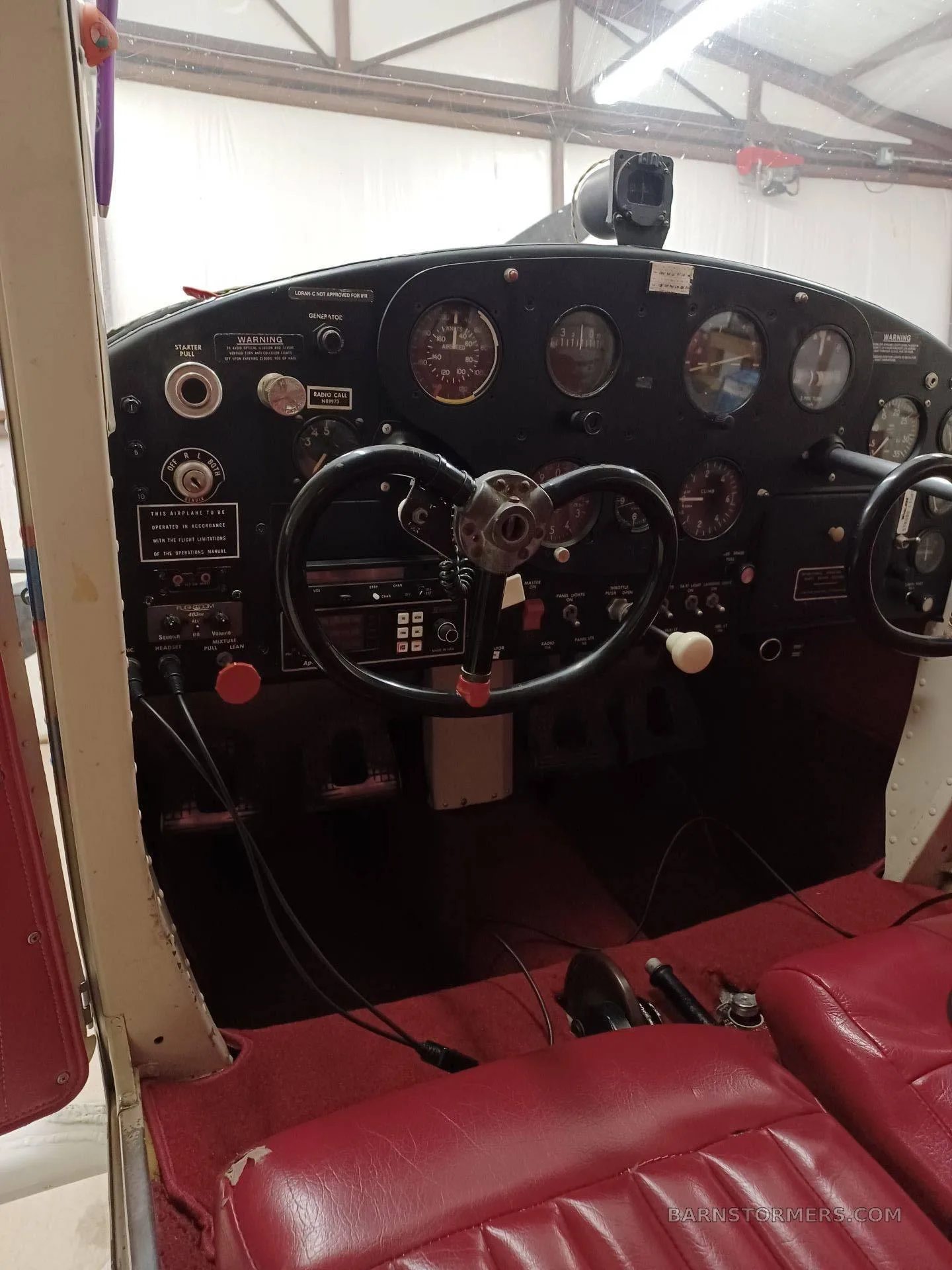 The cockpit of a small aircraft with a black instrument panel, dual yokes, and red upholstered seats.